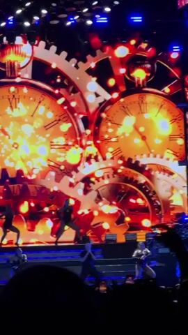 A live concert performance is depicted. The stage features a large LED screen displaying a dynamic visual of oversized clocks with Roman numerals and interlocking gears, predominantly in shades of orange and yellow, accented with sparkling lights. Several dancers in dark, form-fitting costumes are actively performing choreography on a raised platform in front of the screen. In the foreground, the silhouettes of audience members are visible, holding up phones to record the event. The lighting is theatrical and dramatic, illuminating the stage and performers.