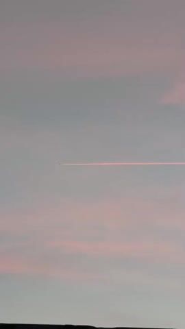 A small airplane leaves a pink contrail across a pale pink and gray sky. The airplane is visible as a tiny dark silhouette against the bright contrail, which stretches horizontally from left to right across the frame. The bottom edge of the frame shows a dark, silhouetted landscape feature, possibly hills or buildings. The overall impression is of a serene, atmospheric scene at dawn or dusk.
