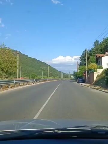 This is a first-person perspective video taken from inside a vehicle traveling along a paved road. The road has a white line dividing the lanes. To the left of the road, there is a metal guardrail, and beyond that, a grassy area and then a tree-covered hillside. A few houses are visible nestled at the base of the hill. The sky is a clear blue with a few scattered white clouds. The sun is shining, indicating daytime. There are utility poles with wires running along the left side of the road. The vehicle appears to be moving at a moderate speed.