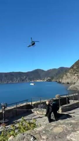 A helicopter is seen flying in a bright blue sky. Suspended by a long cable, the helicopter is carrying a large white package over the sea. The body of water is large and blue, with land visible in the background. The terrain in the background is hilly and covered with vegetation. In the foreground, there is a stone wall and a pair of people standing behind it. One of them is wearing a backpack. Also visible in the foreground is a coin-operated viewing scope.
