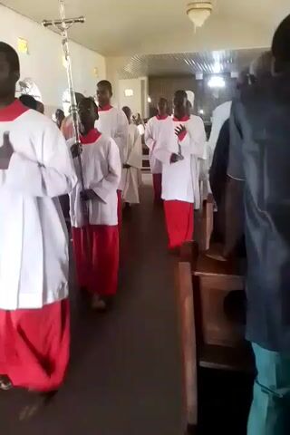 A group of people wearing white robes with red underskirts are walking down the aisle of a church. One person in the middle is carrying a chalice. Others in the group hold a staff. The church has wooden pews and some stained glass windows. Some members of the congregation are visible seated or standing near the pews. The scene suggests a religious procession or service.