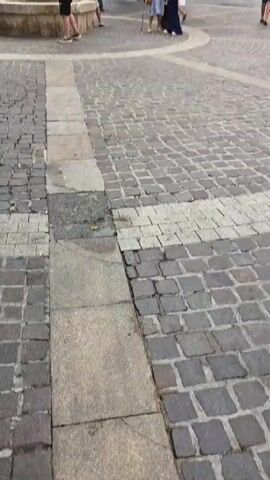 The video shows a bustling outdoor scene in what appears to be a town square or street in Sainte-Maxime, France. The primary subject is the vibrant and colorful canopy of hanging ribbons of various colors that stretches across the overhead view, creating a festive and celebratory atmosphere.

In the foreground, the ground is paved with cobblestones, with a large stone fountain on the left side. Several people are visible, including a man holding a child near the fountain, and other individuals, mostly adults, walking through the square. Some are dressed casually, suggesting it's daytime and likely pleasant weather. A woman with a cane is seen walking, indicating a mix of ages and abilities among the pedestrians.

In the background, there are buildings, including a "SPAR" grocery store with its distinctive green logo, suggesting this is a commercial area within the town. Other people are seen browsing or walking in front of the shops and along the street that recedes into the distance. The overall impression is one of a lively community space, possibly during a local festival or market day, given the elaborate ribbon decoration. The bright colors and the presence of many people evoke a sense of joy and activity. The time of day appears to be daytime, likely late morning or afternoon, with bright lighting.