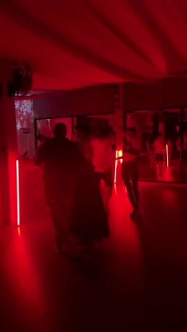 The video shows a group of people dancing in a room bathed in red light. The room has a mirrored wall and a white fabric ceiling. There are vertical red lights. Most people are paired up and dancing. A projection screen is visible in the background. A fan hangs from the ceiling.