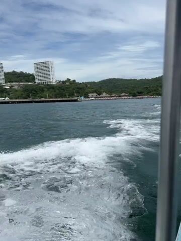 The video captures a view from a moving boat, with white foamy wake visible in the immediate foreground. Dark blue-green water extends towards a coastline featuring a green, tree-covered hill. Two prominent multi-story white buildings are situated on the hill. Below the buildings, a pier or dock structure is visible along the water's edge. To the right of the hill, several smaller structures with conical or thatched roofs line a beach area. The sky is partly cloudy with visible blue patches.