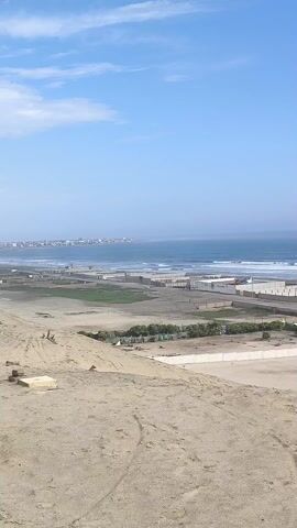 The video presents a high-angle, panning view of a coastal landscape under a clear blue sky. The foreground features a sandy dune area, which transitions into a flat expanse with several rectangular concrete structures or enclosures. Some of these structures contain patches of vegetation. A road runs parallel to the beach, and a single white vehicle is visible moving along it. 

Beyond the structures lies the ocean, with gentle waves rolling towards the shore. In the distance, a faint outline of a town or city is visible along the coastline.

The video pans to the right to reveal a large, rectangular body of water enclosed by concrete and stone structures. A bridge-like feature is visible beneath the water's surface. The overall environment appears to be a mix of natural and man-made elements in a coastal setting.