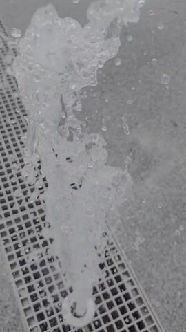 A close-up video shows a jet of water erupting from a drain in a textured, gray surface. The water sprays upwards and outwards, forming a foamy, dynamic plume that breaks apart into smaller droplets. The drain is covered by a white, perforated grate. The video is short and focuses entirely on the water feature.
