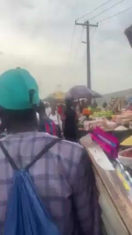 The video shows a busy outdoor market. Numerous people walk through the market, looking at goods displayed on tables and under umbrellas. The tables appear to hold fruits and vegetables. An electrical pole and power lines are visible overhead. The sky is overcast.
