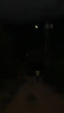 This media file captures a person walking down a dirt path at night, illuminated by a single streetlamp. The setting appears to be rural or undeveloped, with the dark surroundings suggesting a lack of urban sprawl. The streetlamp casts a harsh, bright light on the path, creating stark shadows. The person, seen from behind, is wearing a light-colored shirt. Their movement suggests a journey or a return home. The overall mood is one of solitude and quiet. The time of day is clearly night, with no other light sources visible except for the streetlamp. No weather details can be discerned, but the darkness implies a clear night. There is no visible text in the media file. The location context of San Sebastián, Venezuela, suggests a potentially remote area where such a scene might be common.
