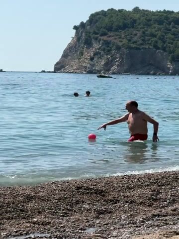 A man in red swim trunks is in shallow water on a pebbly beach, reaching for a pink object in the water. Two other people are swimming further out in the blue ocean. In the background, a large, green, tree-covered hill rises out of the sea. The sky is clear and blue. The man then tosses the pink object into the air. Another man in a striped shirt stands in the water, facing away from the camera, and watches the object fly. Several small islands are visible in the distance.