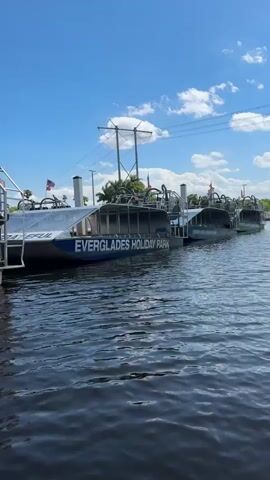 The video shows a row of airboats docked in a body of water. The airboats are labeled "Everglades Holiday Park." The sky is blue with some clouds. There are power lines overhead.