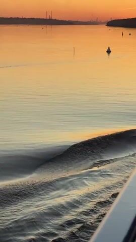 A low-angle shot shows calm water with ripples and a wake caused by a boat moving forward from the right. The water reflects the orange and yellow hues of a sunrise or sunset in the distance. A line of trees with structures is visible on the horizon. A white buoy floats on the water. The scene is viewed from the side of a boat.