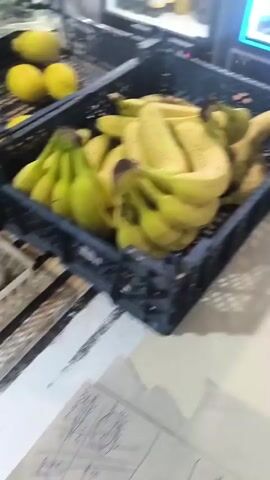 The video opens with a blurry shot of a store aisle with shelves stocked with various products. The camera then focuses on a young woman with dark hair, wearing a light purple t-shirt. She is seen in a store setting, with shelves of products in the background. The woman is looking at the camera, sticking her tongue out playfully, and then waves. The video is shot from a low angle, possibly using a phone.