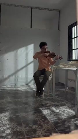 A person is playing a violin while seated on a white chair. The person is wearing an orange shirt, dark pants, and glasses. They are holding the violin under their chin and drawing the bow across the strings. The background is a plain white wall with shadows cast on it. There is a table to the right of the person with some fabric on it. The floor is tiled. The lighting is dim, with a spotlight effect on the wall.