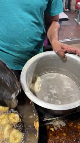 A street vendor wearing a teal shirt is seen frying small, round balls of batter in a large, circular pan filled with oil. He uses a large, slotted spoon to move the fritters around and ensure even cooking. A metal pot containing more batter sits next to the frying pan, and the vendor is shown using his hand to drop the batter into the hot oil. The scene appears to take place outdoors, possibly at a food stall or market.