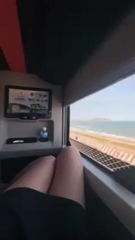 The video presents a view from inside a compartment with a person's legs visible. The person is wearing black shorts and their legs are in the foreground, resting in front of the window. Outside the window is a clear view of a beach with light-colored sand and blue ocean.  A barrier runs parallel to the beach between the viewer and the water.  In the far distance, a small mountain or large hill can be seen. To the left of the window is a small screen displaying various images and a QR code. Below the screen, there's a small tray holding a bottle of water and another unidentified object. The ceiling of the compartment is dark, with a strip of red visible on the left side.  The lighting suggests it is daytime.