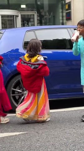 The video shows a street scene with several people and a blue car. In the foreground, a young girl wearing a red jacket over a colorful, traditional Indian dress is standing with her back to the camera. To her left, a woman in a bright pink outfit is visible. To the girl's right, a young boy in a light blue outfit is holding a pink bottle. Another girl, also in traditional Indian attire, is partially visible to the left of the boy. A blue car is parked or driving slowly beside them, with a person in the driver's seat. The background shows a building with large windows and some signage. The ground is asphalt, suggesting a street or sidewalk. The overall atmosphere appears to be a public outdoor setting.