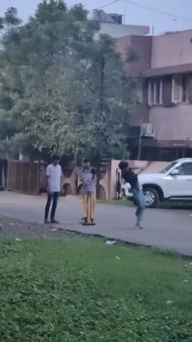 A group of people are playing cricket in an outdoor setting. A person on a motorcycle is seen in the background. The scene appears to be in a residential area with buildings and trees visible.