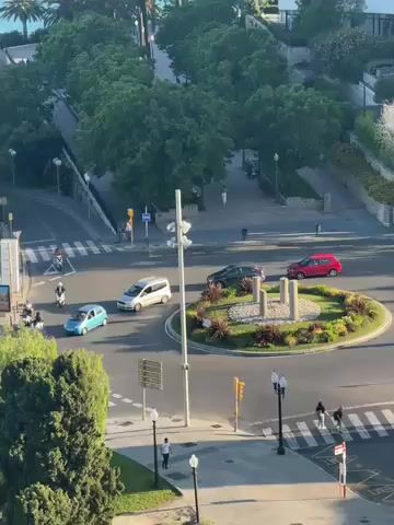 The video presents an elevated, panoramic view of a bustling urban scene in Tarragona, Spain, under clear, sunny skies, likely during morning or late afternoon.

The primary focus is a multi-lane road system featuring a prominent roundabout. Various vehicles are in motion: a red hatchback, a light blue compact car, a white van, and a dark SUV are seen navigating the roundabout or nearby intersections. Several motorcycles and scooters, one with a rider in a red jacket, are also visible. Pedestrians dot the sidewalks and crosswalks, indicating active foot traffic.

In the foreground, three tall flagpoles stand proudly, displaying two Spanish national flags and one Catalan flag. The streets are lined with lush green trees, and the central island of the roundabout is landscaped with greenery and a stone structure. Streetlights and traffic signals are also visible, managing the flow of traffic.

As the camera pans upwards and to the left, the stunning blue expanse of the Mediterranean Sea emerges in the background, with the coastline marked by verdant hillsides dense with trees, including numerous palm trees. A winding coastal road is visible along the cliffs, with a few distant cars. A small building with a red-tiled roof is nestled near the coast. Below the main road, a public green space is discernible, featuring a children's playground with red equipment and shaded pergolas.

The scene captures a dynamic coastal city setting with a blend of urban activity and natural beauty. No discernible text is visible in the video.