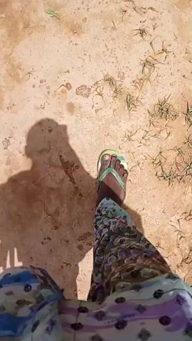 The video shows a first-person perspective of someone walking on dry, cracked earth. The person is wearing patterned pants and green flip-flops. Their shadow is visible on the ground to their left. Sparse, dry grass is scattered across the ground. The camera angle is angled downwards, focusing on the person's feet and legs as they move forward.