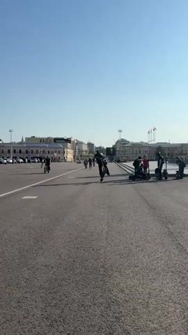 The video shows an outdoor urban environment with a wide asphalt plaza. Several people are present, most of whom are riding electric scooters or bicycles. In the foreground, a person on a scooter is performing a wheelie, with another person on a scooter standing nearby. Further in the background, more individuals are seen riding scooters and bicycles, and some are walking. The scene is set against a backdrop of buildings, some of which appear to be historical or governmental in style. The sky is clear and blue, suggesting a sunny day. The overall impression is of a public space being used for recreation and transportation.
