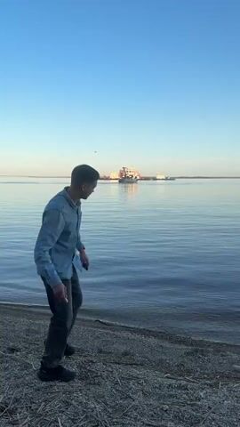A young man stands on the shore of a large body of water and throws a rock into the water. The man is wearing a light-colored button-down shirt, dark pants, and dark boots. He is facing the water and has his right arm raised, ready to throw. The rock is visible in mid-air, just after being released from his hand. The water is calm and blue, with a few ripples where the rock has entered. In the distance, a large barge is visible on the water. The sky is clear and blue. The shore is covered with small rocks and driftwood. The video is shot from a medium distance and is static.