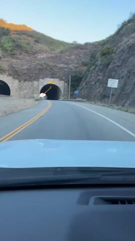 The video shows a first-person view from inside a vehicle approaching and entering a tunnel. The road ahead is paved with double yellow lines indicating no passing. On either side of the road are guardrails. The tunnel is made of stone and has an arched entrance. Yellow warning signs are visible on the sides of the road. Inside the tunnel, there are lights illuminating the pathway. The camera appears to be mounted on the dashboard of the vehicle. The lighting suggests it is either early morning or late afternoon.