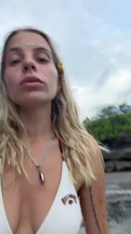 The video shows a fair-skinned woman with long blonde hair standing near the ocean. She is wearing a white bikini top, a silver chain with a purple pendant, and a flower in her hair. She is looking at the camera, and the ocean and rocky coastline are visible in the background. There are other people and vegetation visible nearby. The sky is cloudy.