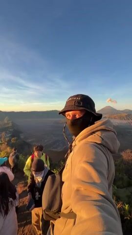 The video is a first-person perspective selfie video with a group of people in the background. The person recording is wearing a black bucket hat, a black face mask, and a tan hooded sweatshirt. They are standing on a grassy slope with a panoramic view of volcanic mountains and a vast, barren landscape under a bright blue sky with wispy clouds. The sun appears to be rising or setting, casting a warm glow on the scene. In the background, several other individuals are visible, also appearing to be on a hike or at a scenic viewpoint. Some are wearing backpacks and casual clothing. The overall environment suggests an outdoor adventure or a visit to a natural landmark.