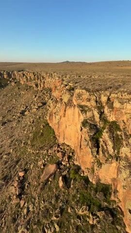 The video captures a tranquil hot air balloon flight over the rugged, arid landscape of Yeşilhisar, Turkey. The scene unfolds at sunrise or early morning, characterized by warm golden light and long shadows stretching across the terrain.

The view is from a hot air balloon, initially looking down at a dramatic, sheer rock face that forms the edge of a deep canyon or plateau. The cliff sides are composed of layered, light-brown rock, interspersed with scattered boulders and sparse, scrubby green vegetation. The ground below the cliffs is equally rocky and uneven.

Throughout the video, the distinct whoosh of the hot air balloon's burner can be heard periodically. At the 0:01 mark, the clear shadow of the camera's hot air balloon becomes visible on the flat, dry plateau just beyond the cliff edge.

As the camera slowly pans across the landscape, it reveals the extensive length of the cliff line, curving into the distance. Several other colorful hot air balloons—white, yellow, and red with partially visible logos—are seen floating gracefully in the clear, bright blue sky in the background, also navigating the unique topography. The sky is entirely cloudless, emphasizing the expansive and serene atmosphere of the flight.
