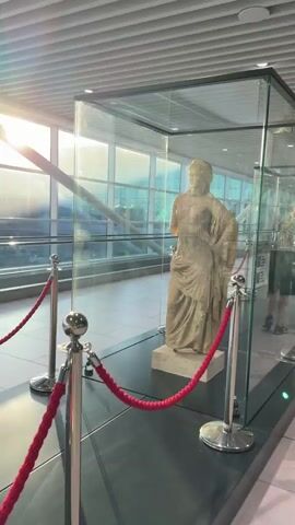 The video shows a static display of sculptures in glass cases. The first sculpture is a standing female figure, partially visible through the glass. The second sculpture, more clearly visible, is a group of figures with a horse, appearing to be a chariot scene. The sculptures are displayed in a well-lit indoor environment, possibly an airport or museum, with large windows showing daylight and an escalator in the background. Red velvet ropes and stanchions are used to keep viewers at a distance from the displays. A person is briefly visible in the background, walking past the display.