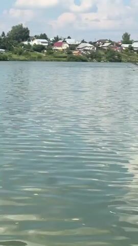 The video shows a lake on a sunny day. In the foreground, the water is rippling and reflects the sky and the surrounding trees. In the middle ground, several people are swimming in the lake. Some are standing in the water, while others are actively swimming. In the background, a wakeboarding ramp is visible, with a person on a wakeboard in motion. There is also a red inflatable boat with people in it. The shoreline is lined with trees and houses. Further down the beach, there are sunbathers and a small building with an umbrella. In the distance, the sky is partly cloudy. The overall scene depicts a recreational area with various water activities taking place.