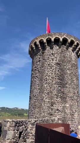 The video shows a tall stone tower with a red flag on top against a blue sky. The tower is made of rough, dark stones and has an ornamental top. The camera pans from the tower to a stone wall and then shows a landscape with buildings, trees, and mountains in the distance. The sky is blue with scattered clouds. There are people visible in the background.