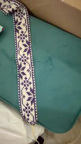 The video pans across three beaded woven bands. The first band, on the left, is purple and white with a snowflake-like pattern. The second band, in the middle, is white, red, and gray with a repeating starburst pattern. The third band, on the right, is dark red, white, and black with a diamond and snowflake pattern. The bands are lying on a teal surface, possibly a cushion, inside a cardboard box. A hand with bright pink nail polish briefly enters the frame at the top, near the second band.