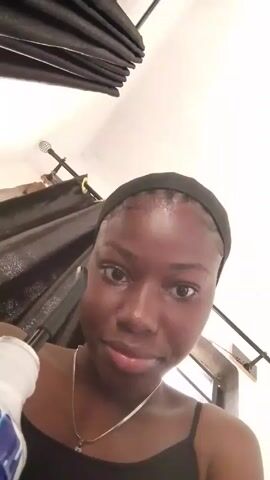 A young Black woman with a black headband, a silver necklace, and a black tank top is seen in a selfie-style video. She is holding a white bottle and a black wand-like object. She pouts her lips. She is in a room with white walls and black curtains. Loud music is playing.