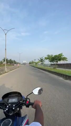 This video captures a first-person perspective of someone riding a motorcycle on a road. The rider's hands are visible on the handlebars, and the motorcycle's dashboard shows a speed of 54, then 55. The road appears to be a highway with multiple lanes, bordered by a curb and a grassy area. On the right side of the road, there are several small, ornamental trees and a fence. Further down the road, a large billboard with the text "JOA" is visible. On the left side of the road, there is a barrier, more trees, and some buildings in the distance. The sky is clear and blue with some scattered clouds. The overall environment suggests a suburban or semi-urban setting.