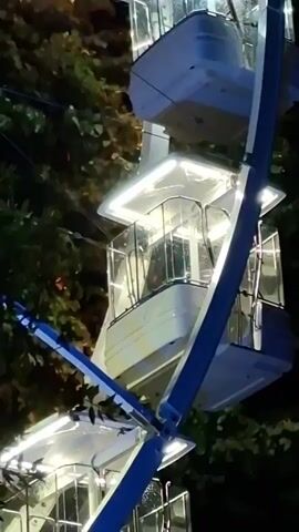 The video shows a close-up, slightly shaky, vertical shot of a Ferris wheel at night. The wheel is illuminated with white lights that shine brightly, highlighting the cabins which are a light color with clear glass windows. The spokes of the wheel are visible, and some appear to be changing color, shifting from red to purple. Dark green foliage from trees is visible in the background, contrasting with the illuminated wheel. The cabins appear to be empty.