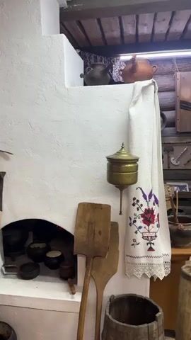 The video shows the interior of a room that appears to be a historical exhibit or a museum display. The room is constructed with log walls and a wooden ceiling. In the foreground, there's a large white stove or oven with wooden paddles leaning against it. To the right, a white embroidered cloth hangs from the stove. Various objects are displayed throughout the room, including pottery, old kitchen tools, furniture like tables and a cabinet, and traditional clothing. There is a hanging cradle in the center of the room. A woman dressed in traditional folk attire, including a red headscarf, is seated on a chair to the right of the cradle. The overall impression is of a preserved historical domestic scene, likely depicting life in a traditional rural setting.