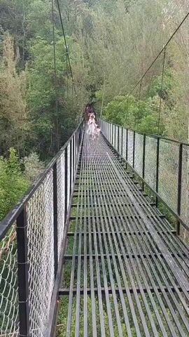 The video shows a long, metal suspension bridge spanning a lush, green forest. The bridge's surface is composed of parallel metal bars, with safety fencing made of chain-link on either side. The railings are black.  A group of people can be seen walking towards the end of the bridge, away from the camera. The background is filled with dense trees.