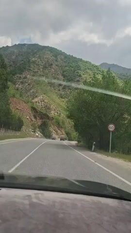 A vehicle travels on a two-lane road through a mountainous landscape. The road is paved with white lines marking the lanes. The surrounding terrain features lush, green hills with trees and some rocky areas. The sky above is cloudy. Two other vehicles are visible ahead on the road. A tall sign is visible on the right shoulder of the road. The camera is mounted inside the moving vehicle, providing the view from the driver's perspective.