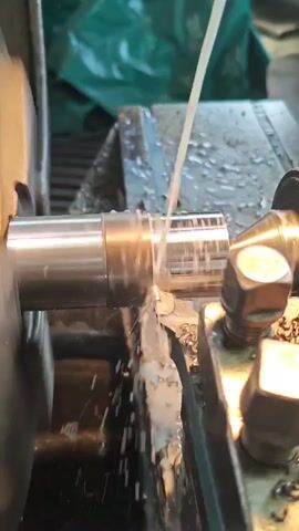 A close-up shot shows a metal rod being machined on a lathe. A cutting tool is in contact with the rod, removing material as it spins. A stream of coolant is being sprayed onto the cutting area to lubricate and cool the tool and workpiece. Metal shavings are visible being ejected from the cutting area. The background is blurred but appears to be an industrial setting with green material draped in the upper left.