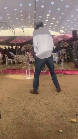 The video shows an outdoor event, likely a wedding reception, taking place under a large white tent decorated with patterned lights. Several people are visible, including a man in a white shirt and jeans dancing, a man in a blue robe, and a young woman with braided hair wearing a white shirt and gray pants. A video camera is visible, suggesting the event is being recorded. The ground appears to be dirt and grass. There are tables and chairs set up under the tent, and floral decorations are scattered around.