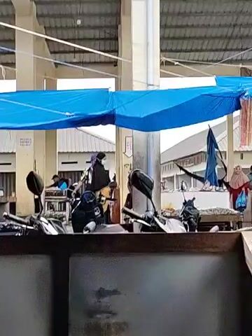 The video shows a marketplace scene in what appears to be Southeast Asia. The market is under a covered structure with white pillars and a metal roof. A blue tarp provides additional shade. Several motorcycles are parked in the foreground, obscuring part of the view. People are milling about, some dressed in traditional Muslim clothing. There are market stalls visible in the background where vendors seem to be selling goods. A few people are standing at the stalls, possibly making purchases. The overall environment suggests a typical, bustling marketplace.