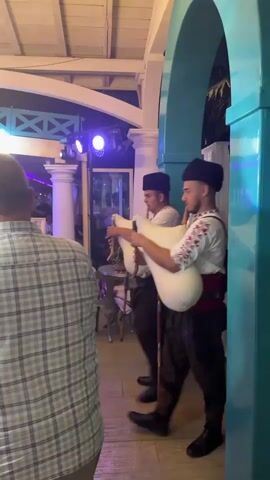 Two young men dressed in traditional clothing, consisting of black hats, white shirts with embroidered sleeves, red sashes, and dark pants, play bagpipes. They stand amidst a dimly lit venue with a wooden ceiling and white architectural elements. In the background, other people are visible, some appearing to dance or socialize. There are also purple lights illuminating the space, and a chandelier hangs from the ceiling. A ring light is set up to the left of the frame. A man in a plaid shirt is partially visible in the foreground, facing away from the camera. The bagpipes appear to be made of white material.