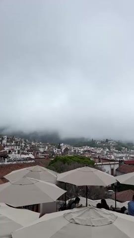 A rooftop cafe scene with a view of a misty town and a church. Several large beige umbrellas are visible in the foreground, some partially obscuring the view. Underneath the umbrellas, several people are seated at tables, appearing to be dining or socializing. They are mostly out of focus and their faces are not clearly visible. Beyond the cafe, a town with red-tiled roofs and white buildings cascades down a hillside. The town is partially obscured by fog and clouds. A large, ornate church with two towers and scaffolding on one tower is prominent in the middle ground, to the right. The sky is overcast and the overall atmosphere is muted due to the weather.