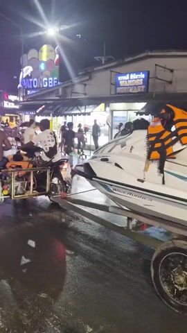 This video captures a nighttime street scene. A jet ski on a trailer is being towed by a motorcycle. The trailer and motorcycle are on a wet road, reflecting the lights from the surrounding environment. In the background, there are buildings with illuminated signs, including one that reads "PATONG BEACH" and another that says "WANDER MART." Several people are visible, some on other motorcycles and some walking. The overall atmosphere appears to be that of a busy tourist area at night, possibly after rain.