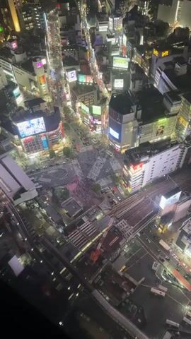 The video shows a high-angle view of Shibuya Crossing in Tokyo at night. The crossing is filled with pedestrians walking in all directions. Cars are stopped at the surrounding streets waiting for the light to change. There are many large buildings with illuminated billboards and signs surrounding the intersection. A train station and rail lines are visible in the foreground. The overall lighting is bright due to the city lights and illuminated signs. The scene is bustling with activity.