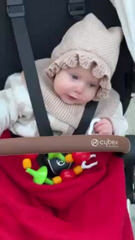 A baby sits in a black Cybex stroller, secured by black straps. The baby wears a beige knitted hat with a ruffled edge and a matching knitted collar. They are dressed in a white and beige patterned outfit and are sucking on a colorful teething ring with red, yellow, and green elements. A red blanket is draped over the stroller's safety bar. The stroller is being pushed along a sidewalk. The background is blurred.