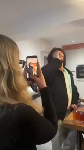 A woman is filming a video of a man drinking from a glass. The woman has long blonde wavy hair and is wearing a black shirt. The man is wearing a black jacket over a light blue and white shirt. He has curly dark hair. The man is standing at a bar. On the bar are bottles of alcohol. In the background, there are kitchen cabinets and an oven. The man tastes the drink and then speaks. The woman films the man with her phone.