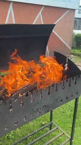 The video shows a close-up of a metal grill with wood burning inside. The flames are bright orange and engulf the wood. The grill is positioned on a metal stand, and in the background, there is a building with an orange and white patterned wall and green grass. The focus is on the fire and the grill.