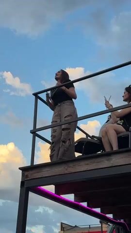 A woman stands on a raised platform, looking up at the sky and holding a phone in her left hand. She is wearing a black t-shirt, distressed denim pants, and her hair is down. To her right, another woman is seated, also holding a phone and looking to the side. The platform has a metal railing and wooden flooring, and is supported by metal beams. In the background, the sky is filled with dramatic clouds, illuminated by the setting sun. Below the platform, there are buildings and a stage area, with some graffiti visible. The overall atmosphere appears to be an outdoor event or gathering.